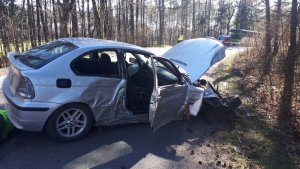 Prawa strona samochodu bmw, uszkodzenia na przednich i tylnych drzwiach wgniecenia, maska podniesiona do góry, koło przednie prawe wyrwane, leży na boku, w tle za samochodem policjant w kamizelce odblaskowej.