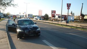 Samochód bmw z rozbitą przednią szybą, za nim policjanci na drodze przeprowadzają oględziny.