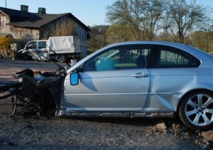 Na zdjęciu widoczny lewy bok samochodu osobowego, przód samochodu całkowicie rozbity, w tle dalej widoczny volkswagen, który wjechał w ogrodzenie posesji.