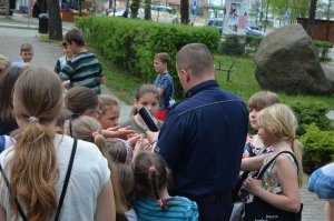 policjant przed budynkiem komendy rozdaje dzieciom uczestniczącym w spotkaniu odblaskowe opaski