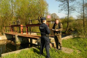 policjant wraz z  funkcjonariuszem straży rybackiej legitymują wędkarza przy zbiorniku wodnym przy lesie