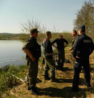 policjant wraz z  funkcjonariuszami straży rybackiej legitymują wędkarzy przy zbiorniku wodnym przy lesie, rozmawiają z nimi