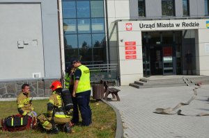 Policjanci znajdują się przed budynkiem UM w Redzie i pomagają strażakowi przy ewakuowanej kobiecie