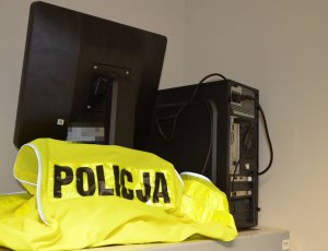 Na zdjęciu znajduje się zabezpieczony komputer, jest ustawiony przodem do ściany, przed nim leży kamizelka odblaskowa z napisem POLICJA.