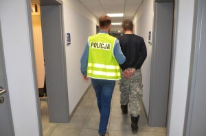 Policjant w odblaskowej kamizelce prowadzi zatrzymanego korytarzem komendy