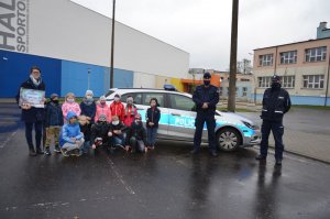 Zdjęcie grupowe klasy pierwszej z policjantami po prawej stronie na tle radiowozu i hali sportowej przy szkole podstawowej nr 5 w Wejherowie