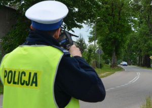 Policjant w żółtej odblaskowej kamizelce stoi tyłem i mierzy urządzeniem trucam prędkość nadjeżdżających z oddali samochodów