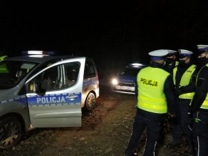 Policjanci stoją na ciemnej leśnej drodze, po lewej stronie zdjęcie znajduje się radiowóz