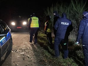 jadący radiowóz po lewej stronie a po prawej idzie grupa policjantów ciemną leśna drogą