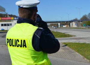 Na zdjęciu widzimy plecy i głowę policjanta stojącego tyłem do robiącego zdjęcie. Policjant stoi z miernikiem prędkości trucam przyłożonym do oka i mierzy prędkość nadjeżdżającego samochodu z oddali