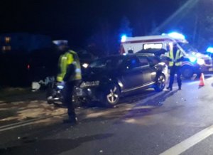 Zdjęcie ciemne, widać światła błyskowe w kolorze niebieskim, na pierwszym planie policjanci przed rozbitym samochodem w kolorze czarnym, w tle widoczna karetka