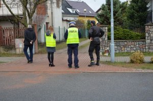 Na chodniku stoi czterech członków komisji. Stoją obróceni tyłem do zdjęcia, oglądają miejsce zdarzenia i infrastrukturę drogową. Na zdjęciu od lewej wysoki mężczyzna w czarnej kurtce, następnie po jego prawej stronie stoi kobieta policjantka ubrana w ubiór cywilny, na kurtce ma założoną żółtą kamizelkę odblaskową, obok niej po prawej stoi umundurowany policjant ruchu drogowego ( w białej czapce) , a następnie obok niego po prawej stoi strażak ubrany w czarny mundur (moro) z czapką z daszkiem. Na chodniku niedaleko przy strażaku zostały ustawione znicze.