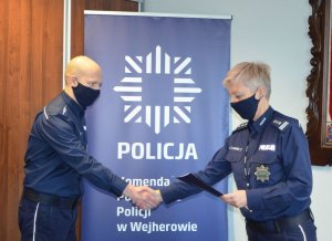 Na pierwszym planie po prawej pani Komendant gratuluje policjantowi, który stoi po lewej stronie zdjęcia. Oboje są ustawieni bokiem do osoby fotografującej. Ubrani są w granatowe mundury. W tle za nimi po środku widoczne jest tło - baner z policyjną gwiazdą i napisem Komenda Powiatowa Policji w Wejherowie.