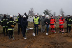 Na zdjęciu stoi grupa 12 osób, w tym wójt, policjanci, strażacy, ratownicy medyczni i inni goście. Stoją na terenie działki z łopatami wbitymi w ziemię.