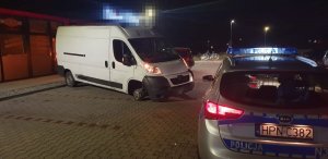 Na parkingu przy budynku po lewej stoi biały samochód peugeot boxer, bez przedniej opony, na zniszczonej feldze, po prawej na pierwszym planie zajęcia widać tył oznakowanego radiowozu.