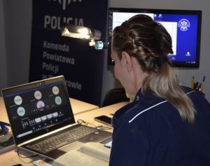 Policjantka siedzi przy biurku, na którym otwarty jest komputer laptop. Na ekranie widać okienka z inicjałami dzieci uczestniczących w lekcji on-line.