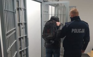 Policjanci prowadzi zatrzymanego mężczyznę przy pomocy dźwignie transportowej na ramię. Wprowadza go do celi która znajduje się po lewej stronie.