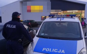 Policjant stoi po lewej stronie samochodu, a przed nim znajduje się market spożywczy, nazwa sklepu jest niewidoczna. Po prawej stronie w oddali widać wchodzących klientów do sklepu.