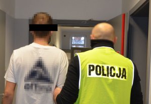 Policjant po prawej stronie w kamizelce odblaskowej z napisem POLICJA prowadzi zatrzymanego mężczyznę do celi