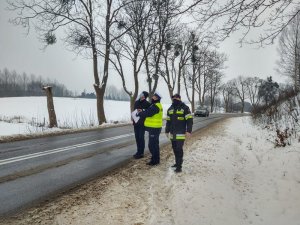 Policjant, strażak i przedstawiciel komisji stoją na poboczu drogi  i sprawdzają infrastrukturę.