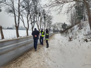 Policjant, strażak i przedstawiciel komisji stoją na poboczu drogi  i sprawdzają infrastrukturę.