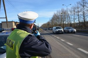 Policjant stoi tyłem do fotografującego, wykonuje pomiar prędkości nadjeżdżających z przeciwka samochodów.