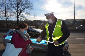 Policjant wręcza opaskę odblaskową kobiecie. W tle za nimi widoczny jest policyjny radiowóz.