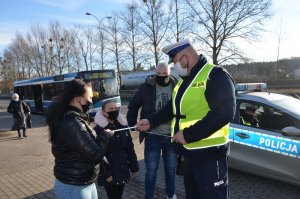 Policjant wręcza opaski odblaskowe mężczyźnie, dziecku i kobiecie, w tle za nimi stoi radiowóz, a w oddali autobus.