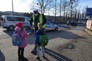 Policjant wręcza odblaski dwójce dzieci, które idą z plecakami. W tle za policjantem widoczny jest radiowóz.