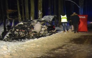 Zdjęcie wykonane nocą, widoczny jest czarny samochód z otwartą klapą bagażnika, przód rozbity, samochód stoi przed drzewem, w tle policjant w odblaskowej kamizelce wykonuje czynności służbowe