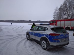 Radiowóz stoi przed jeziorem, za nim w tle wóz straży pożarnej, na zamarzniętym jeziorze stoi człowiek i lekko widać jedynie wystający dach samochodu