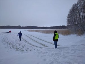 Policjant stoi przed zamarzniętym jeziorem, za nim inny mężczyzna idzie w kierunku brzegu, w tle widać klęczącego mężczyznę który zagląda pod wodę na zamarzniętym jeziorze w miejscu gdzie wpadł samochód