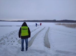 Policjant stoi przed zamarzniętym jeziorem, za nim widać dwóch innych mężczyzn na jeziorze w pobliżu miejsca gdzie wpadł samochód