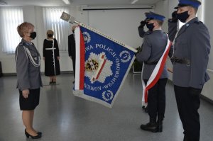 Pani Komendant żegna się ze sztandarem, stoi na wprost sztandaru i trzymającego go policjanta, po którego obu stronach stoją dwaj policjanci, w tle widać kobietę ubraną w czarną sukienkę z białymi aplikacjami oraz mężczyznę w czarnym garniturze