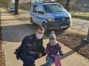 Policjant pozuje do zdjęcia z dziewczynką na rowerku biegowym, przykucnął w pewnej odległości od dziewczynki, w tle widoczny jest radiowóz.