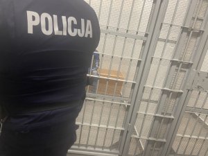 Policjant zamyka drzwi pomieszczenia dla osób zatrzymanych, przez kratę w drzwiach częściowo widać zatrzymanego mężczyznę.