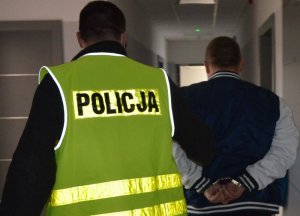 Policjant prowadzi zatrzymanego . Zatrzymany ma kajdanki założone na nadgarstki, policjant trzyma go za przedramię, widać ich plecy