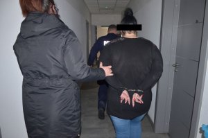 Policjantka prowadzi zatrzymaną kobietę, jest prowadzona w kajdankach założonych na ręce trzymane z tyłu, przed nimi idzie policjant w granatowym mundurze