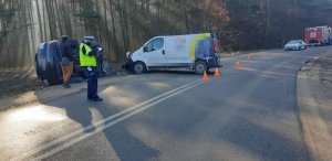 Policjant stoi przed rozbitymi samochodami, wykonuje oględziny. Samochód po lewej stronie leży na dachu.