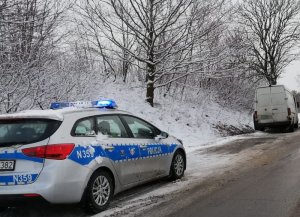 Na pierwszym planie na poboczu stoi osobowy zaparkowany radiowóz, następnie trochę dalej jest biały samochód który uderzył w drzewo, w tle widoczny śnieg na poboczu oraz na drodze