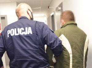 Policjant prowadzi zatrzymanego mężczyznę, który ubrany jest w kurtkę w kolorze khaki z białymi pionowymi pasami