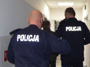 Dwóch policjantów prowadzi zatrzymanego, nie widać koloru jego kurtki