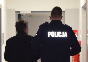 Policjant prowadzi zatrzymanego, który jest ubrany w czarną kurtkę