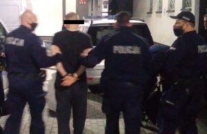 Dwóch policjantów prowadzi zatrzymanego mężczyznę, trzeci stoi obok i asekuruje czynności