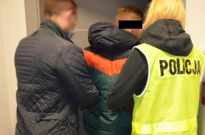 Od prawej policjantka ubrana w kamizelkę odblaskową, po lewej policjant w granatowej kurtce, po środku między nimi zatrzymany mężczyzna, który jest przez nich prowadzony do wyjścia.