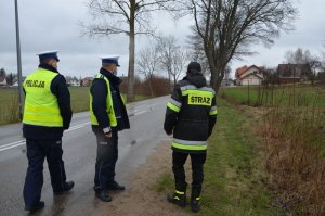 Na drodze, na jej skraju stoją dwaj policjanci a koło nich po prawej stoi strażak, w tle widoczne jest drzewo - miejsce wypadku śmiertelnego.