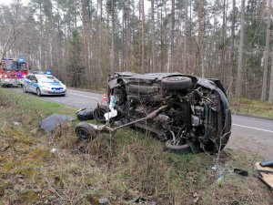 Na pierwszym planie widoczny jest volkswagen leżący  na boku, za nim w tle stoi policyjny radiowóz, a dalej wóz straży pożarnej.