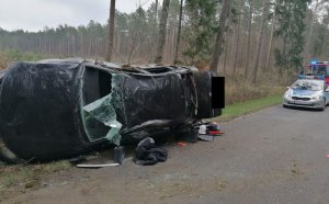 Na pierwszym planie widać rozbity leżący na boku samochód, widoczne są wgniecenia uszkodzenia karoserii i szyby,