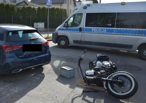 Na jezdni leży na boku motocykl, tuż obok po lewej stoi samochód z którym się zderzył - mercedes, a po prawej w tle policyjny radiowóz.