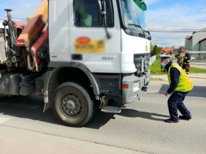 Samochód ciężarowy stoi na jezdni, policjant przed samochodem pochyla się i ogląda przód pojazdu.